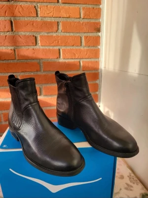 Vagabond svarta chelsea boots i skinn 38 - Säljer ett par klassiska svarta chelsea boots i skinn med rund tå och låg klack. Skorna har elastiska paneler på sidorna och dragkedja på insidan för enkel påtagning. Perfekta för dig som gillar stilrena och tidlösa boots.