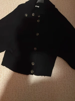 Svart kofta med knappar. - Säljer en svart kofta med snygga stora knappar framtill. Koftan har lång ärm, hög krage och råa kanter nertill för en edgy look. Perfekt att slänga över en t-shirt eller topp för extra stilpoäng. Akryl. Säljs på fler sidor.