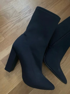 Svarta stövletter med klack och spetsig tå - Säljer ett par svarta stövletter med hög klack och spetsig tå. Skorna har dragkedja på insidan och är tillverkade i ett stretchigt syntetmaterial som sitter snyggt runt benet. Perfekta för dig som vill ha en stilren och edgy look.