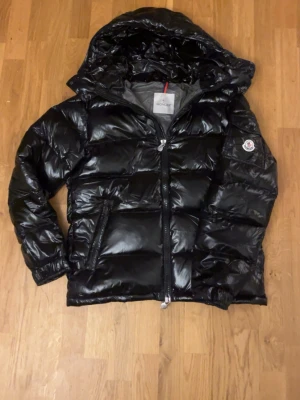 Svart Moncler pufferjacka med huva - Säljer en svart pufferjacka från Moncler med glansig finish och huva. Jackan har dragkedja framtill, två sidofickor med dragkedja och klassisk Moncler-logga på ärmen. Insidan har ett coolt serietryck och jackan är perfekt för kalla dagar.