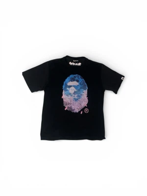 Svart BAPE t-shirt med blått tryck - Svart t-shirt från BAPE med ikoniskt ape-head tryck i blått och lila på bröstet. Klassisk passform med korta ärmar och rund hals. Liten BAPE-logga på ärmen och tryck i nacken. Perfekt för dig som gillar streetwear och vill sticka ut.