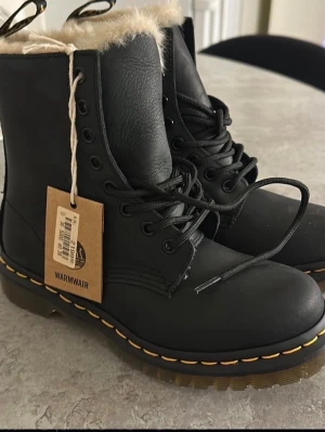 Svarta Dr. Martens boots med foder - Säljer ett par svarta Dr. Martens boots med varmt beige foder och klassisk gul söm runt sulan. Skorna har snörning, rund tå och är tillverkade i skinn. Perfekta för kalla dagar och ger en riktigt snygg edge till din outfit.