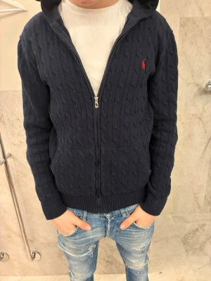 Marinblå kofta från Polo Ralph Lauren - Säljer en marinblå kabelstickad hoodie från Polo Ralph Lauren med dragkedja och huva. Tröjan har långa ärmar, fickor framtill och den klassiska röda loggan på bröstet. Storleken är XL 18-20 så den passar XS/S, Hör av er vid frågor!