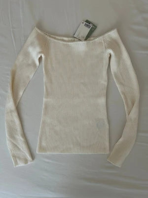 Offshoulder ribbad topp med pärlor - Säljer en superfin offshoulder topp i cremevitt med ribbad struktur. Toppen har långa ärmar och en snygg pärldetalj längs urringningen som ger en extra touch. Materialet känns mjukt och stretchigt. Helt ny och oanvänd med prislapp på.