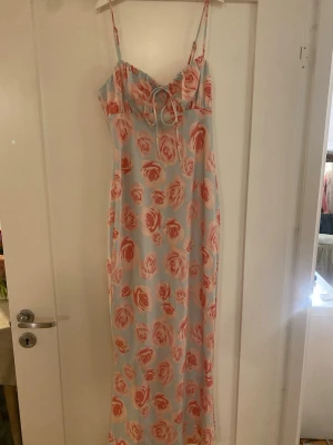 Lång blommig klänning med snörning - Supersöt långklänning med ljusblå bas och stora rosa blommor. Klänningen har tunna axelband, v-ringning och en söt snörningsdetalj framtill. Perfekt för sommarens alla tillfällen och ger en romantisk vibe.