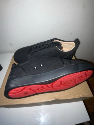 Christian Louboutin svarta sneakers - Säljer ett par svarta sneakers från Christian Louboutin i mocka med ikoniska röda sulor och broderad logga bak. Skorna har snörning och rund tå, samt exklusiva detaljer och medföljande dustbag och certifikat.