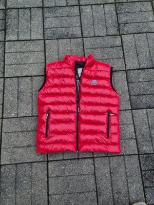 Moncler - Säljer en röd dunväst från Moncler med glansig finish och ståkrage. Västen har två dragkedjeförsedda fickor framtill och tydlig Moncler-logga på bröstet. Perfekt för lager-på-lager och snygg till både hoodie och t-shirt.