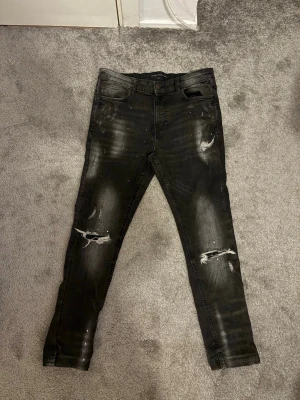 Jeans  - Säljer ett par svarta skinny jeans med slitna detaljer och hål på knäna. Jeansen har en tvättad look och är i stretchigt material för skön passform. Perfekta för dig som gillar en edgy stil. Byxorna är från Jd (supply and demand)