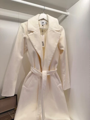 Vit kappa från JDY med bälte - Elegant kappa från JDY i en krämvit nyans. Stilren modell med klassisk krage, långa ärmar och praktiska fickor. Den medföljande knytningen i midjan ger en fin siluett och gör att kappan sitter riktigt bra. Aldrig använd och i nyskick med lappen kvar. JDY är ett eget märke som tillhör samma modeföretag som ONLY (båda är en del av BESTSELLER-gruppen). Hör av dig om du vill se hur den sitter på! 