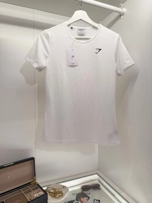 Vit T-Shirt - Gymshark - Vit T-shirt från Gymshark med svart logga på bröstet. T-shirten har rund halsringning och är tillverkad i ett mjukt och stretchigt material. Enkel och stilren design. Aldrig använd och i nyskick med lappen kvar. 