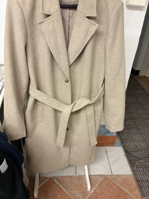 Beige kappa från VILA med bälte - Snygg beige kappa från VILA i klassisk modell med bälte i midjan och stora slag. Kappan har knappar framtill, två fickor och är helfodrad. Perfekt för höst och vinter när du vill ha en stilren look. Den är i knlängd. Super fint skick extremt sparsamr anvönd bara lite nopprigt under armhålorna men det går nog att raka av. Nypris 900kr.