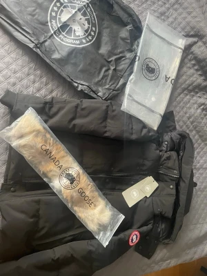 Svart dunjacka från Canada Goose - Den visar storlek L men passar folk som har S och M. Svart Canada Goose dunjacka med klassisk patch på ärmen och avtagbar pälskrage. Jackan har dragkedja, knappar och flera praktiska fickor. Perfekt för kalla vinterdagar och riktigt snygg streetstil.