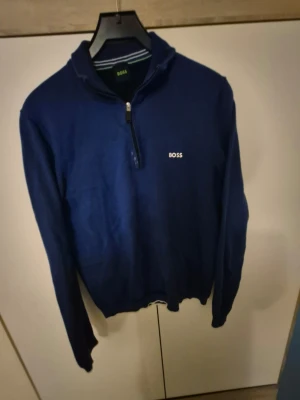 Blå half zip tröja från BOSS - Snygg blå tröja från BOSS med half zip och vita detaljer vid muddar och krage. Tröjan har lång ärm och ribbade avslut. Perfekt för dig som vill ha en stilren och sportig look. Broderad BOSS-logga på bröstet och randiga detaljer i kragen.