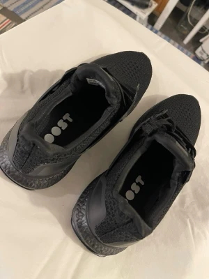 Svarta Adidas Ultraboost sneakers - Säljer ett par helsvarta Adidas Ultraboost sneakers med stickad ovandel och klassiska tre ränder på sidan. Skorna har snörning, platt sula och BOOST-dämpning för extra komfort. Perfekta för dig som vill ha både stil och funktion i vardagen.