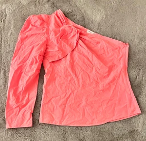 Korallrosa oneshoulder topp Loavies - Säljer en korallrosa oneshoulder topp från Loavies med lång ärm på ena sidan och snygg puffdetalj vid axeln. Toppen har en cool asymmetrisk design och är gjord i ett lätt, slätt material som känns skönt mot huden.