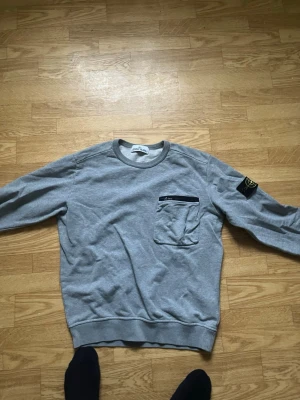 Grå sweatshirt från Stone Island - Säljer en grå sweatshirt från Stone Island med klassisk patch på ärmen och en bröstficka med svart dragkedja. Mycket bra skick. Tröjan har inge hål eller skador