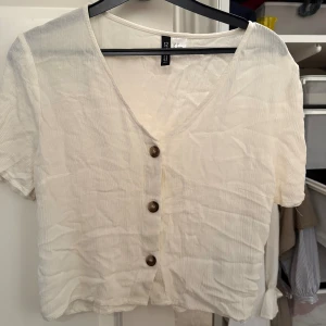 Vit kortärmad blus från H&M Divided - Säljer en ljus, krämvit blus från H&M Divided i storlek 36. Toppen har korta ärmar, v-ringning och bruna knappar framtill.💗