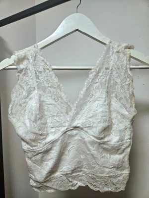 Vit spetsblus med v-ringning - Superfin vit blus i spets med v-ringning både fram och bak. Blusen är ärmlös och har ett blommigt spetsmönster över hela plagget. Perfekt för dig som gillar romantiska detaljer och vill ha något som sticker ut lite extra. Vissa bilder såg den gul ut mot banden men har tagit en närbild så man förstår att det bara är ljuset som gör att det ser ut så! 