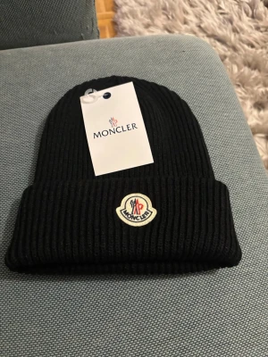 Svart Moncler mössa - Säljer en svart ribbstickad mössa från Moncler med uppvikt kant och klassisk Moncler-logga framtill. Mössan är tillverkad i mjukt material och har en stilren look som passar till många outfits.