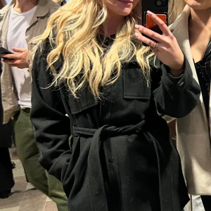 Svart kappa med snörning från Bubbleroom - Stilren svart skjortkappa från Bubbleroom med snöre i midjan och stora bröstfickor. Jackan har klassisk krage, knappar framtill och långa ärmar. Perfekt för dig som vill ha en snygg och tidlös look under höst och vinter. Sparsamt använd.