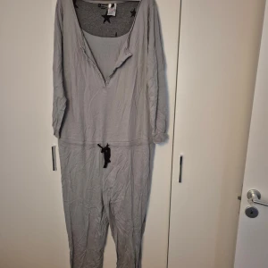 Grå jumpsuit från Rainbow, 2XL - Grå jumpsuit från Rainbow i 100% bomull med långa ärmar, knappar framtill och snörning i midjan. Mjuk och skön, perfekt för chill dagar. Stjärnmönster på insidan av kragen ger en extra touch.