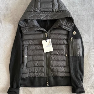Moncler cardigan  - Snygg svart  cardigan från Moncler med stickade ärmar och huva. Jackan har tvåvägsdragkedja, fickor med dragkedja på ärmen och ribbad nederkant. Ikonisk Moncler-logga på vänster ärm. Perfekt för dig som vill ha en cool och modern look.