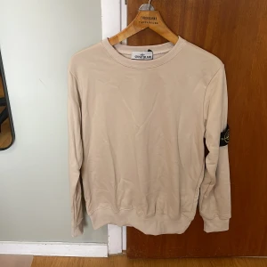 Beige sweatshirt från Stone Island - Säljer en beige sweatshirt från Stone Island med klassisk rund hals och ribbade muddar. Tröjan har den ikoniska Stone Island-loggan på vänster ärm. Perfekt för dig som gillar stilrena och enkla plagg med exklusiv känsla.