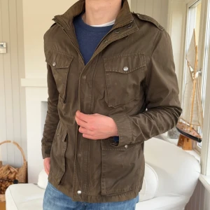 Olivgrön fieldjacket från Woolrich - Säljer en olivgrön fieldjacket från Woolrich med fyra stora fickor framtill och snygga axelklaffar. Jackan har dragkedja och knappar, klassisk krage och är tillverkad i slitstark bomull. Perfekt för dig som gillar funktionell och stilren design.