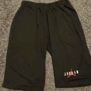 Svarta Jordan shorts med logga - Säljer ett par svarta Jordan shorts med den klassiska Jumpman-loggan i rött och vitt på ena benet. De har elastisk midja och är tillverkade i ett mjukt syntetmaterial, perfekta för sport eller chill. Passformen är loose och längden går ner mot knäna.