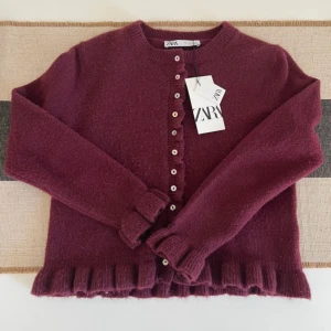 Vinröd kofta med volang från Zara - Supersöt vinröd kofta från Zara med volangkant och pärlemorknappar framtill. Mjuk och fluffig känsla tack vare blandning av polyester, akryl och ull. Rund hals och långa ärmar, perfekt för att addera lite extra stil till din outfit.