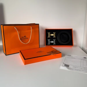 Svart H bälte med H-spänne - Exklusivt svart bälte från H Paris i äkta skinn med präglad logga och subtilt mönster. Levereras med två utbytbara H-spännen i guld och silver. Snyggt och stilrent, perfekt för dig som gillar lyxiga accessoarer och vill sticka ut med klass.