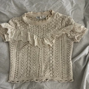 Beige stickad blus med volang från Zara - Superfin beige stickad blus från Zara med korta ärmar och broderad volangdetalj över bröstet. Blusen har ett vackert hålmönster och ribbad halsringning. Perfekt för dig som gillar romantiska och unika detaljer.