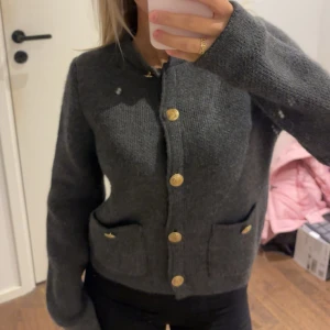 Mörkgrå stickad kofta med guldknappar - Mysig mörkgrå stickad kofta med snygga guldknappar framtill och på fickorna. Koftan har två fickor och en klassisk rak passform med långa ärmar. Perfekt för lager-på-lager och chill höststil. 