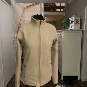 Beige fleecejacka med dragkedja - Snygg beige fleecejacka med hel dragkedja framtill och höga krage. Tröjan har två praktiska fickor med dragkedja och är långärmad. Perfekt för kyliga dagar, att ha under jackan.
