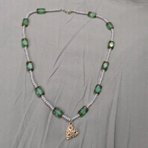 Halsband med gröna pärlor och hjärthänge - Unikt halsband, ca 60cm långt, med rektangulära gröna pärlor och små ljusgrå pärlor. I mitten hänger ett coolt hjärthänge i silverton med hålmönster. Perfekt för dig som gillar färg och detaljer! Lås i metall för enkel användning.