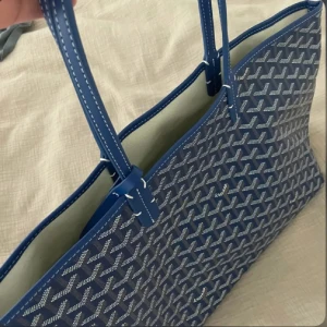 Blå shoppingväska  - Säljer en blå shoppingväska med deras ikoniska mönster i vitt och grått. Väskan har långa handtag och är rymlig, perfekt för att bära med sig allt man behöver. Tillverkad i slitstarkt syntetmaterial med läderdetaljer.