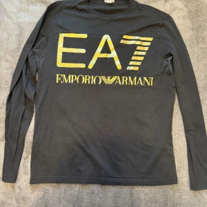 Svart långärmad EA7 Emporio Armani tröja - Svart långärmad tröja från EA7 Emporio Armani med stor, glansig guldfärgad logga och text på bröstet. Rund hals och mjukt bomullsmaterial som känns skönt mot huden. Perfekt för dig som gillar stilrena och lyxiga streetwear-plagg.