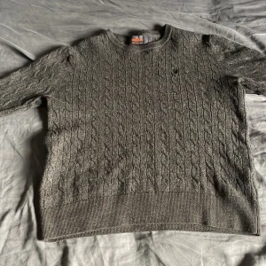 Mörkgrå kabelstickad tröja Morris - Snygg mörkgrå kabelstickad tröja från Morris i 100% merinoull. Tröjan har rund hals, ribbade muddar och ett diskret broderat emblem på bröstet. Perfekt för dig som gillar klassisk stil med en modern touch.