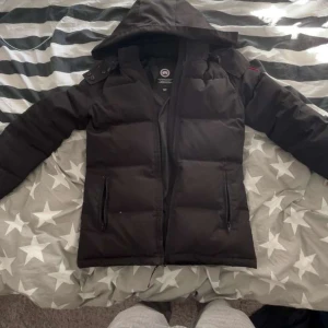 Svart dunjacka Canada Goose M - Svart dunjacka från Canada Goose med huva och klassisk röd/vit logga på ärmen. Jackan har två sidofickor med dragkedja och quiltad design. Perfekt för kalla dagar och har en clean, stilren look som funkar till allt. priset kan såklart diskuteras vid snabb affär.