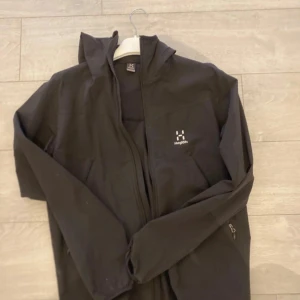 Svart jacka med huva från Haglöfs - Säljer en svart jacka med huva från Haglöfs. Jackan har dragkedja framtill och är långärmad. Perfekt för utomhusaktiviteter med sin stilrena design och funktionalitet.