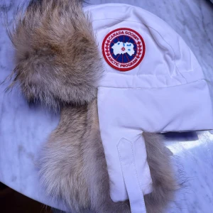 Vit pälsfodrad trappermössa Canada Goose - Säljer en vit trappermössa från Canada Goose med äkta pälsdetaljer runt öron och panna. Mössan har ett broderat märke framtill och är fodrad för extra värme. Perfekt för kalla vinterdagar och har en klassisk, snygg look.