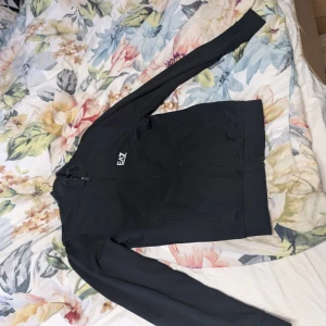 Svart tröja från EA7 Emporio Armani - Svart tröja från EA7 Emporio Armani med stilren design och vit logotyp på bröstet. Tröjab har hel dragkedja framtill, ribbade muddar och lång ärm. Materialet är mjukt och känns som bomull, perfekt för en chill och snygg look.