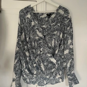 Svartvit omlottblus från Lindex  - Säljer en svartvit omlottblus från Lindex med abstrakt prickigt mönster. Blusen har långa ärmar och en snygg omlottfront