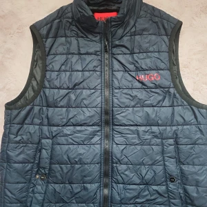 Mörkblå quiltad dunväst från Hugo - Quiltad mörkblå dunväst från Hugo med röd logga på bröstet. Västen har hög krage, dragkedja framtill och två fickor med knappdetaljer. Perfekt för lager-på-lager och stilren look.