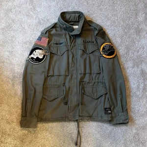 Grön militärjacka från Ralph Lauren - Säljer en militärinspirerad grön jacka från Ralph Lauren Denim & Supply med coola patches på båda ärmarna, bland annat amerikansk flagga och broderade motiv. Jackan har flera fickor, snörning i midjan och dragkedja i halsen. Perfekt för dig som gillar streetstyle. Storlek S-M
