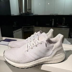  Adidas Ultraboost  - Säljer ett par fräscha vita Adidas Ultraboost sneakers med stickad ovandel och klassiska tre ränder på sidan. Skorna har snörning, platt sula och en riktigt bekväm boost-mellansula. Perfekta för dig som gillar stilrena och sportiga skor.