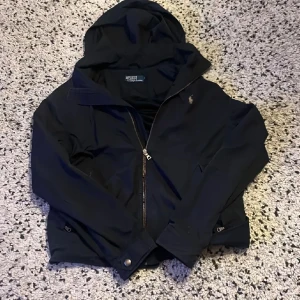 Svart vindjacka från Polo Ralph Lauren - Snygg marinblå windbreaker från Polo Ralph Lauren med huva och dragkedja. Jackan har ett broderat logotypmärke på bröstet och knappar vid ärmslut och nederkant. Perfekt för en stilren look. Jackan har ett bra skick förutom dragkedjan som det fattas några tänder ifrån men funkar oftast annars bra skick.