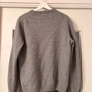 Grå stickad tröja från Lyle & Scott L - Snygg grå stickad tröja från Lyle & Scott med rund hals och deras klassiska gula fågel-logga broderad på bröstet. Tröjan har ribbade muddar vid ärmslut och nederkant, och är långärmad. Perfekt för dig som gillar stilren och enkel design.