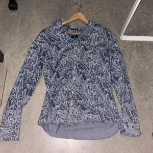 Blå mönstrad skjorta från Gant - Snygg blå skjorta från Gant med vitt bladmönster och klassisk krage. Skjortan har knappar framtill och är långärmad. Modellen är fitted och materialet känns som bomull, perfekt för dig som gillar stilrena men ändå unika plagg.