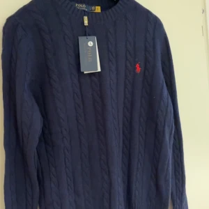Marinblå kabelstickad tröja Polo Ralph Lauren - Kabelstickad marinblå tröja från Polo Ralph Lauren med rund halsringning och klassisk röd broderad logga på bröstet. Tröjan är långärmad och har ribbade muddar vid ärmslut och nederkant. Perfekt för lager-på-lager under kyliga dagar.